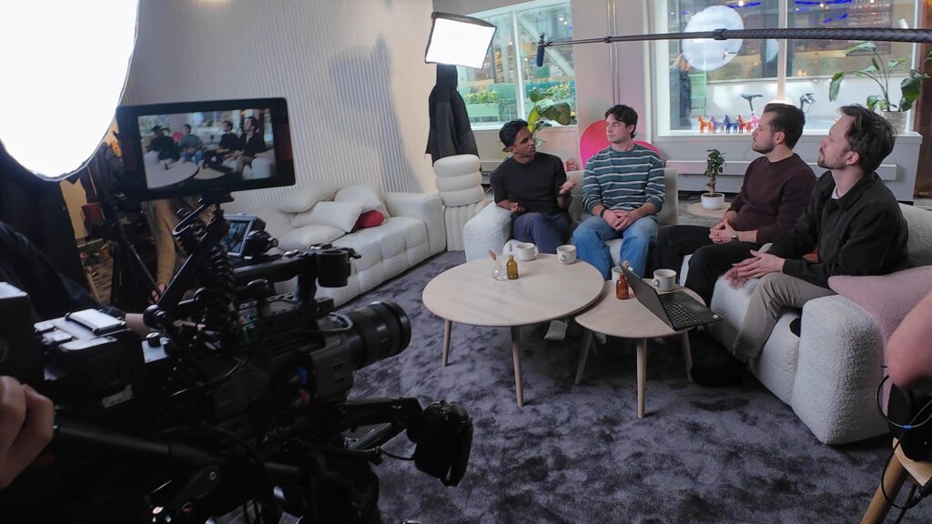 Bakom kulisserna vid videoproduktion med kamera, studioljus och fem personer sittande i soffor under en filmad gruppdiskussion i en modern lounge.