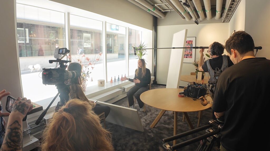 Bakom kulisserna vid intervjuproduktion med kamerateam, bommikrofon, ljusutrustning och en kvinna som talar framför kameran vid stora fönster i en modern kontorsstudio.