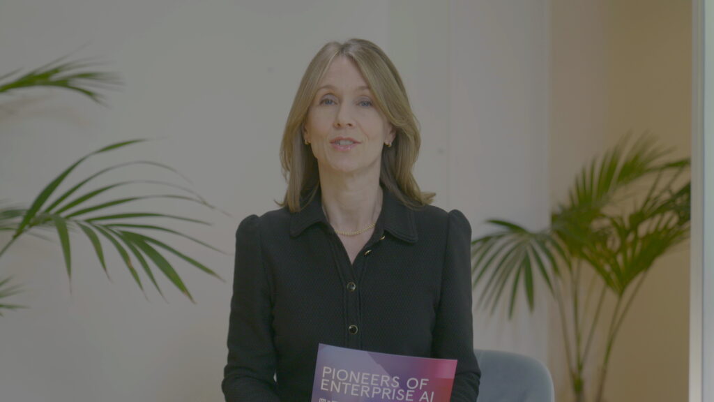 Professionell kvinna i svart klädsel talar framför kameran, håller ett kort med texten ”Pioneers of Enterprise AI”, sittande inomhus med dekorativa växter i en ljus modern miljö.
